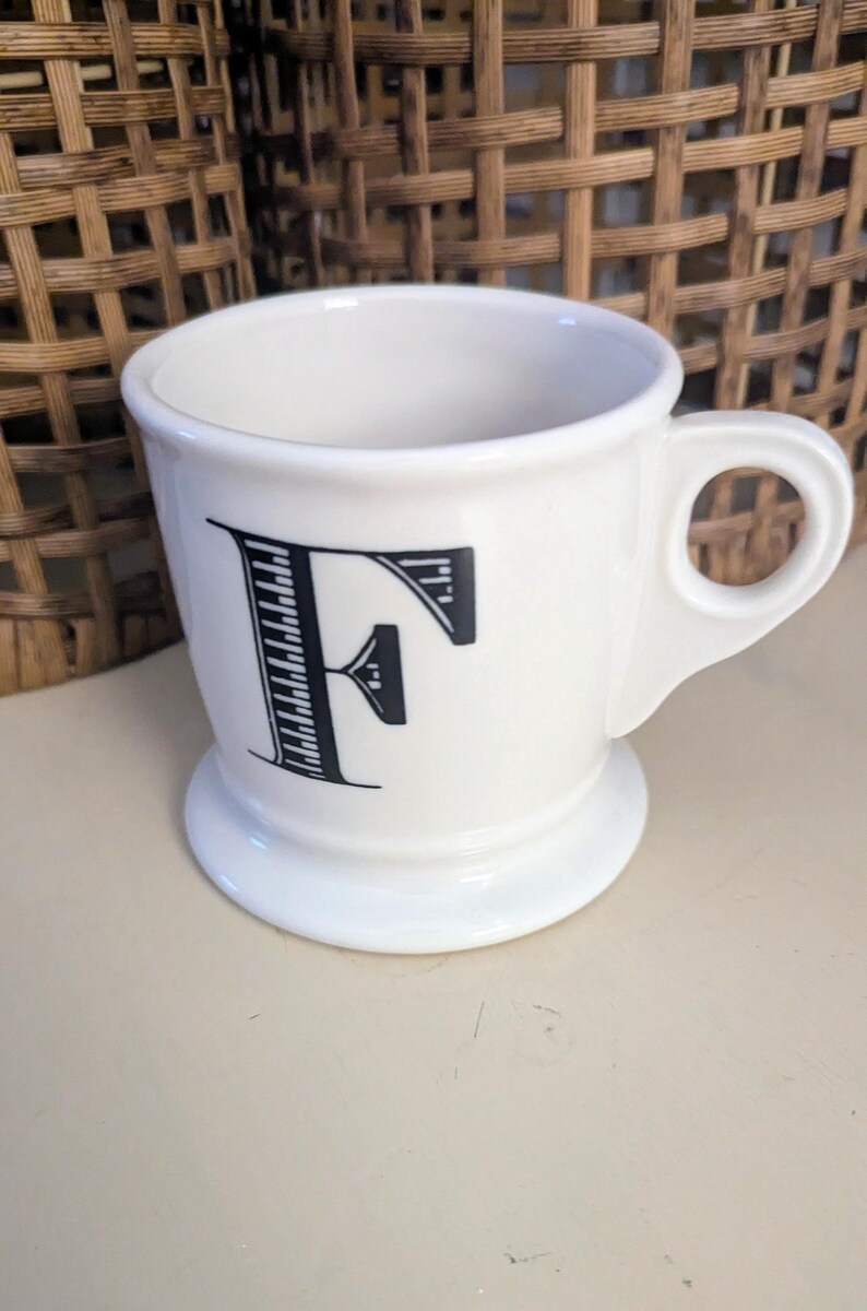 K&ouml;nnte beinhalten: Wei&szlig;e Keramiktasse mit schwarzem Buchstaben "F" auf der Vorderseite. Die Tasse hat einen Henkel und einen Sockel. Im Hintergrund ein Weidenkorb.