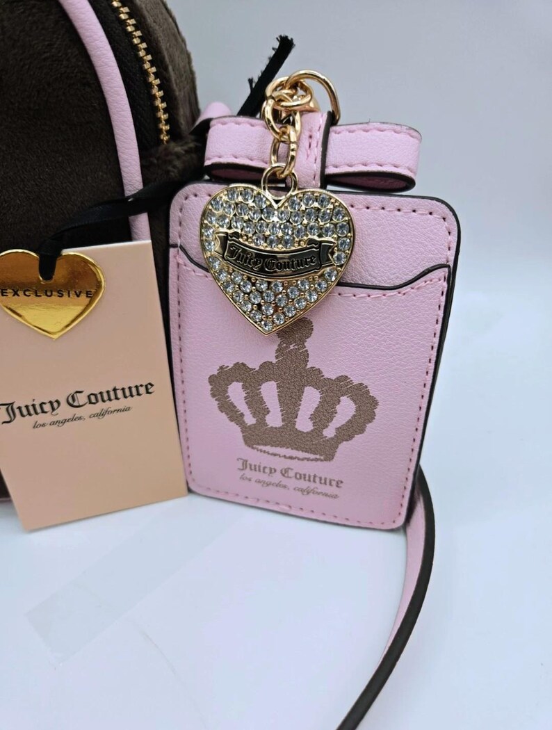 May include: A pink Juicy Couture cardholder with a gold crown design and a heart-shaped charm. The charm is encrusted with rhinestones and features the Juicy Couture logo. A gold heart-shaped tag reads "EXCLUSIVE".