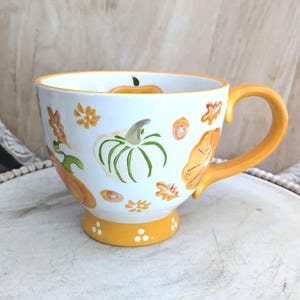 May include: White ceramic mug with orange handle and base, decorated with hand-painted pumpkins, leaves, and flowers. The mug has a wide, rounded shape, perfect for enjoying a warm beverage. A charming fall-themed design.