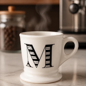 May include: A white ceramic mug with a large black letter "M" on the front. The letter is partially striped. Steam rises from the mug, suggesting a hot beverage. The mug sits on a marble surface.