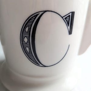 K&ouml;nnte beinhalten: Eine wei&szlig;e Keramiktasse mit einem gro&szlig;en schwarzen Buchstaben "C" auf der Vorderseite. Der Buchstabe ist mit dekorativen Linien und Formen versehen. Die Tasse hat einen Henkel und einen leicht ausgestellten Fu&szlig;.