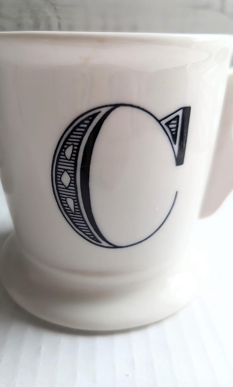 K&ouml;nnte beinhalten: Eine wei&szlig;e Keramiktasse mit einem gro&szlig;en schwarzen Buchstaben "C" auf der Vorderseite. Der Buchstabe ist mit dekorativen Linien und Formen versehen. Die Tasse hat einen Henkel und einen leicht ausgestellten Fu&szlig;.