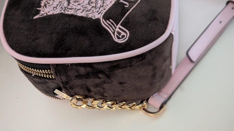 May include: A small, black velvet backpack with a pink trim and strap. The bag features a gold zipper and chain detail. The top of the bag has a pink embroidered design. The bag is a stylish accessory.