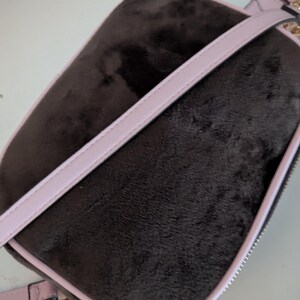May include: A dark brown, rectangular crossbody bag with a soft, velvety texture. The bag features a light pink trim and strap, with a gold-colored zipper and heart-shaped charm. The bag is designed for carrying essentials.