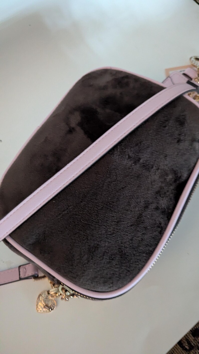 May include: A dark brown, rectangular crossbody bag with a soft, velvety texture. The bag features a light pink trim and strap, with a gold-colored zipper and heart-shaped charm. The bag is designed for carrying essentials.