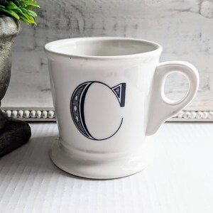 K&ouml;nnte beinhalten: Wei&szlig;er Keramikbecher mit einem gro&szlig;en, stilisierten schwarzen Buchstaben "C" auf der Vorderseite. Der Becher hat einen abgerundeten Boden, einen gebogenen Henkel und ein schlichtes, klassisches Design.