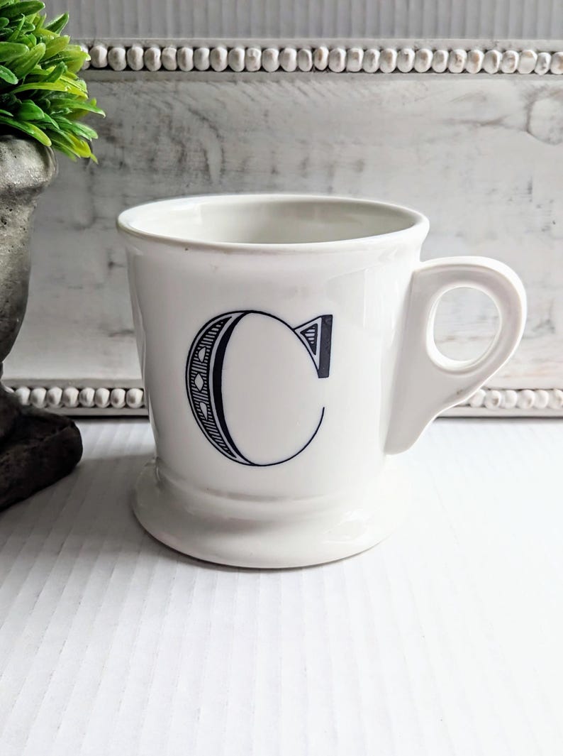 K&ouml;nnte beinhalten: Wei&szlig;er Keramikbecher mit einem gro&szlig;en, stilisierten schwarzen Buchstaben "C" auf der Vorderseite. Der Becher hat einen abgerundeten Boden, einen gebogenen Henkel und ein schlichtes, klassisches Design.