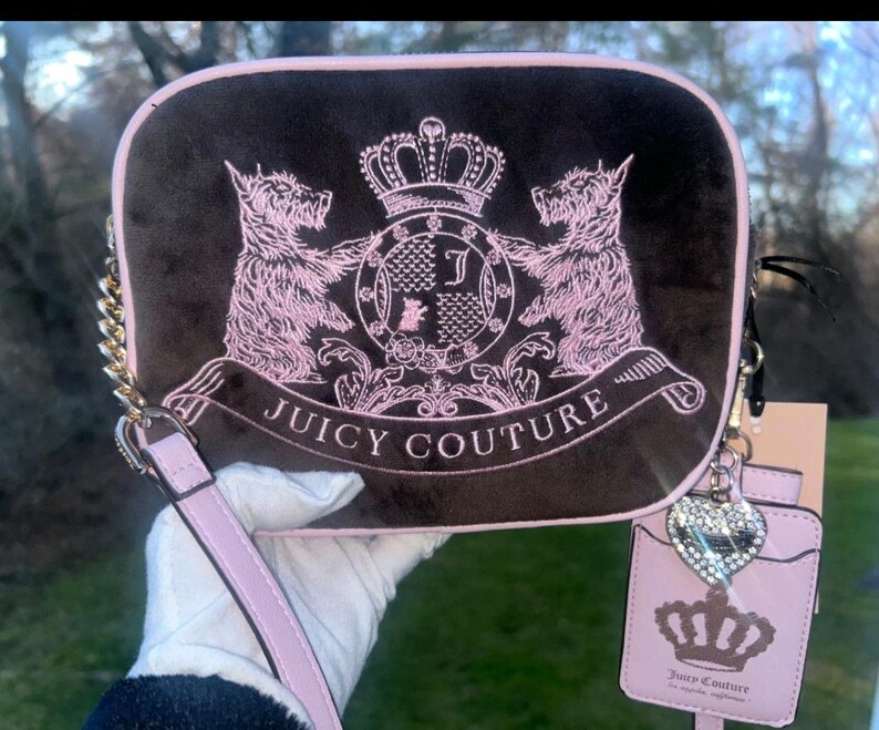 May include: A brown rectangular Juicy Couture handbag with a pink trim, featuring a pink embroidered crest with two dogs, a crown, and the brand name. It has a gold chain strap and a pink wristlet.
