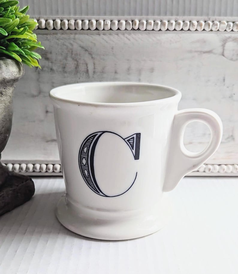 K&ouml;nnte beinhalten: Wei&szlig;e Keramiktasse mit einem gro&szlig;en, stilisierten schwarzen Buchstaben "C" auf der Vorderseite. Die Tasse hat einen abgerundeten Boden und einen Henkel. Der Hintergrund ist eine hellfarbige Oberfl&auml;che.