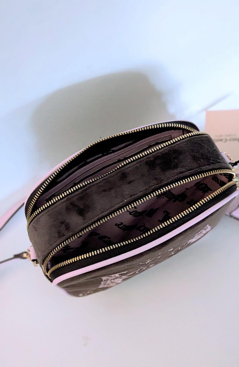 May include: Black and pink crossbody bag with gold zippers. The bag has a rounded shape and features a pink interior lining. The exterior is black with a subtle pattern. The bag has a pink strap.