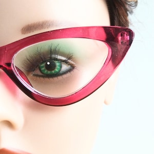 May include: Close-up of a mannequin's face wearing cat-eye glasses. The frames are a vibrant magenta color, contrasting with the green eye shadow and the mannequin's skin tone. The eye has long, dark eyelashes and a bright green iris.