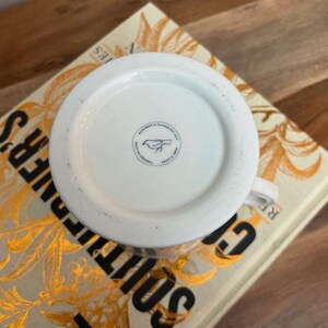 K&ouml;nnte beinhalten: Wei&szlig;er Keramikbecher mit rundem Boden, mit einem kleinen Vogellogo und Text. Der Becher steht auf einem Buch mit goldfarbenen Buchstaben und einem floralen Design. Der Becher ist mit "Microwave & Dishwasher Safe Made in China" beschriftet.