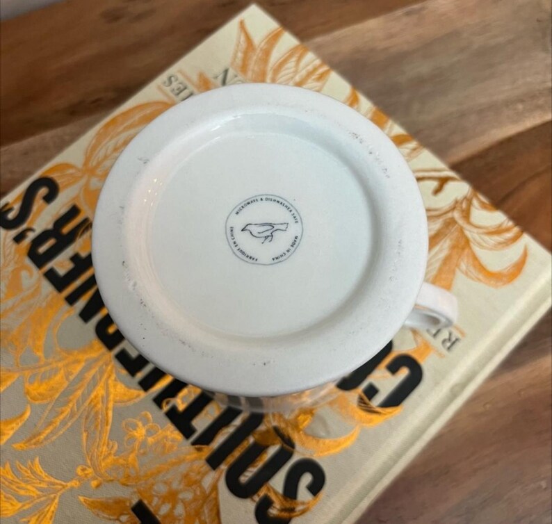 K&ouml;nnte beinhalten: Wei&szlig;er Keramikbecher mit rundem Boden, mit einem kleinen Vogellogo und Text. Der Becher steht auf einem Buch mit goldfarbenen Buchstaben und einem floralen Design. Der Becher ist mit "Microwave & Dishwasher Safe Made in China" beschriftet.