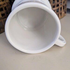 K&ouml;nnte beinhalten: Eine wei&szlig;e Keramiktasse mit Henkel, von oben gesehen. Die Tasse hat eine weite &Ouml;ffnung und einen leicht ausgestellten Rand. Der Innenraum ist glatt und wei&szlig;. Ein kleiner Henkel ist an der Seite angebracht.