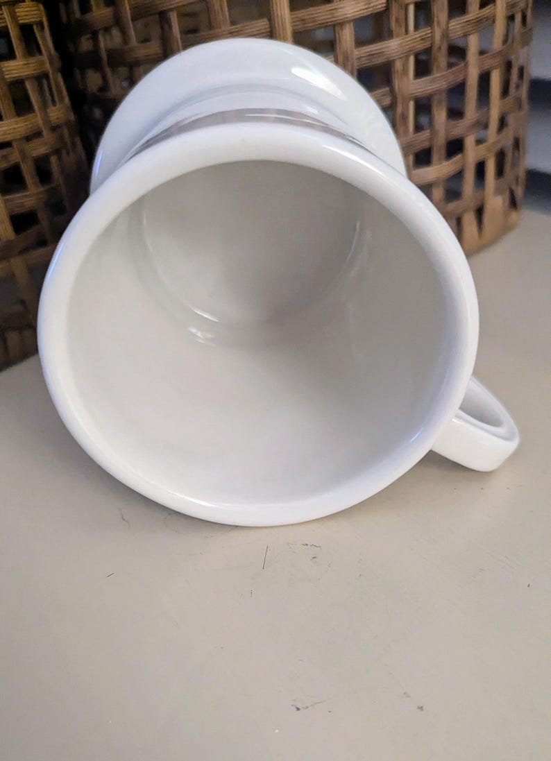 K&ouml;nnte beinhalten: Eine wei&szlig;e Keramiktasse mit Henkel, von oben gesehen. Die Tasse hat eine weite &Ouml;ffnung und einen leicht ausgestellten Rand. Der Innenraum ist glatt und wei&szlig;. Ein kleiner Henkel ist an der Seite angebracht.