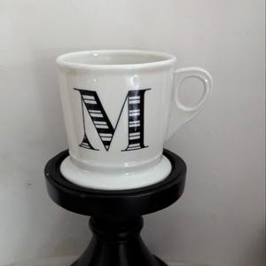 May include: White ceramic mug with a black letter "M" design, set on a black decorative stand. The mug has a handle and the letter is filled with vertical stripes. The stand has a round top and a pedestal base.