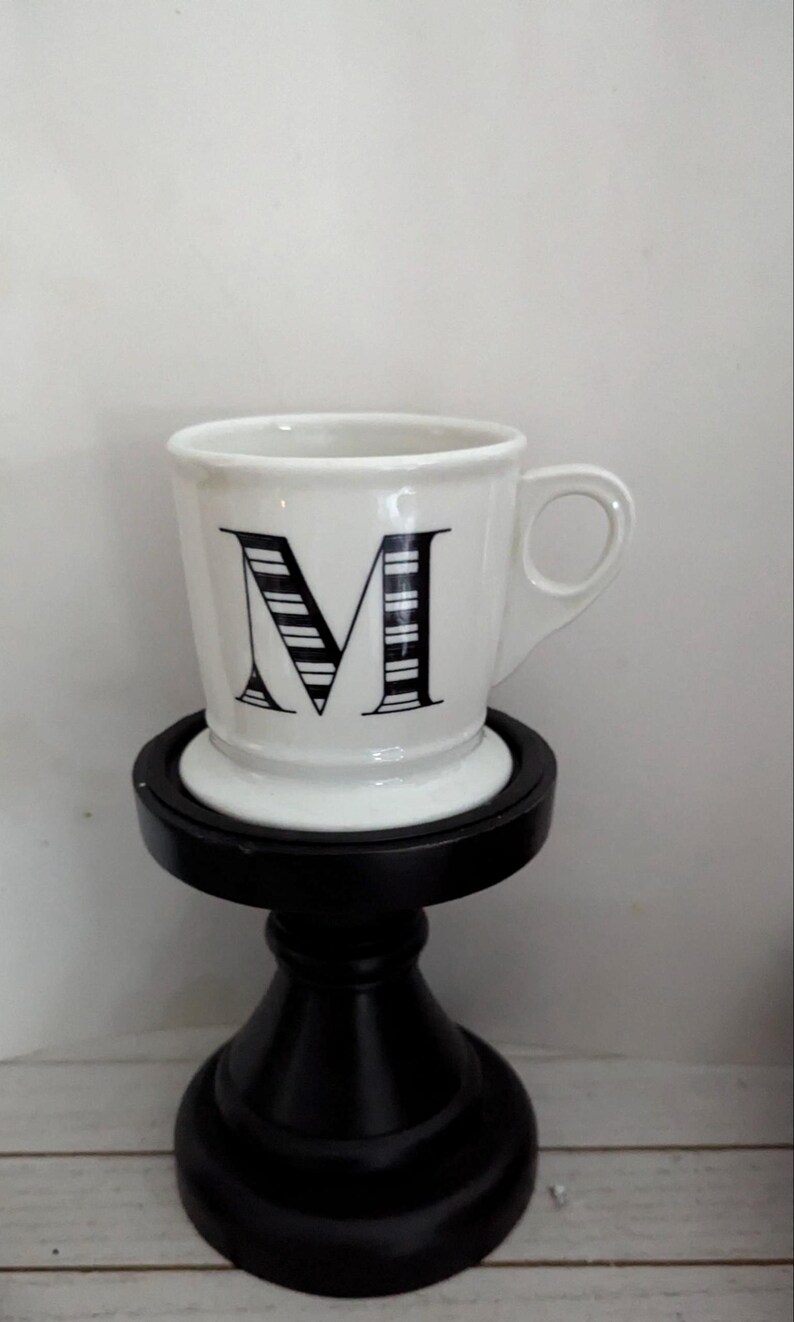 May include: White ceramic mug with a black letter "M" design, set on a black decorative stand. The mug has a handle and the letter is filled with vertical stripes. The stand has a round top and a pedestal base.