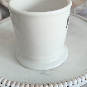 K&ouml;nnte beinhalten: Eine wei&szlig;e Rasierbecher aus Keramik mit ausgestelltem Fu&szlig;, auf einem dekorativen wei&szlig;en Tablett mit Perlenrand. Der Becher hat ein kleines, dunkles Etikett. Der Hintergrund ist eine helle Holzoberfl&auml;che.