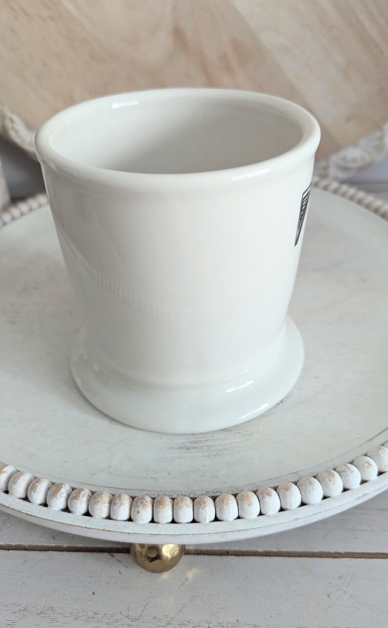 K&ouml;nnte beinhalten: Eine wei&szlig;e Rasierbecher aus Keramik mit ausgestelltem Fu&szlig;, auf einem dekorativen wei&szlig;en Tablett mit Perlenrand. Der Becher hat ein kleines, dunkles Etikett. Der Hintergrund ist eine helle Holzoberfl&auml;che.