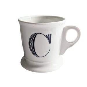 K&ouml;nnte beinhalten: Wei&szlig;e Keramiktasse mit schwarzem Buchstaben "C". Die Tasse hat einen abgerundeten Boden, einen Henkel und ein schlichtes, elegantes Design. Ideal f&uuml;r Kaffee, Tee oder andere Getr&auml;nke.