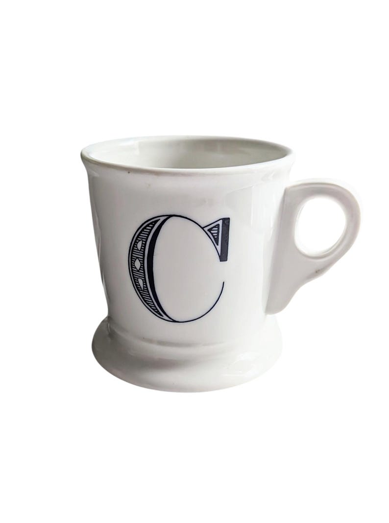 K&ouml;nnte beinhalten: Wei&szlig;e Keramiktasse mit schwarzem Buchstaben "C". Die Tasse hat einen abgerundeten Boden, einen Henkel und ein schlichtes, elegantes Design. Ideal f&uuml;r Kaffee, Tee oder andere Getr&auml;nke.