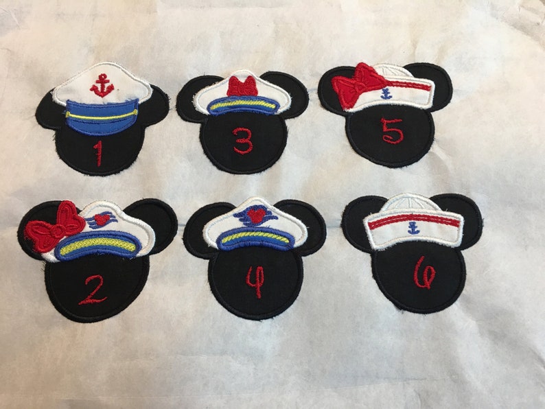 May include: Six black Mickey Mouse head-shaped patches with sailor hat designs. Each patch has a different hat design in white, red, blue, and yellow, with an anchor or heart emblem. Numbers 1-6 are embroidered in red.
