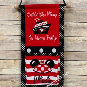 May include: A red and black fabric wall hanging with a pocket. The top section features a white ship with red hearts and the text "Cruisin' With Mickey" and "The North Family". The bottom pocket features a red and white striped fabric with a Minnie Mouse applique. The text "Minnie" is embroidered below the applique. The text "Threadin' Stitches" is embroidered in the bottom right corner.