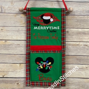 Op de afbeelding: Een groene stoffen wandhanger met een rood-groene geruite rand. Het bovenste gedeelte heeft de woorden "Very Merrytime Cruise" en een Mickey Mouse-ontwerp met een kerstmuts. Het onderste gedeelte heeft een Mickey Mouse-hoofd met kerstverlichting en de naam "Brad".