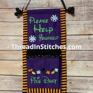 May include: A purple fabric wall hanging with orange and black striped side borders. It features the embroidered text "Please Help Yourself" and "Pixie Dust" in green, along with two circular blue accents and a pocket decorated with small colorful icons.