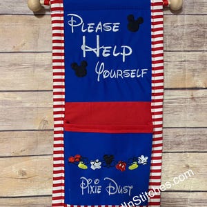 May include: A blue wall hanging with the words "PLEASE HELP YOURSELF" and "Pixie Dust" embroidered in white. Mickey Mouse silhouettes and shoe designs are also embroidered. The hanging has a red pocket and is trimmed with red and white stripes.