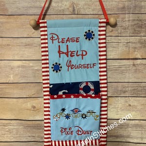 May include: A light blue fabric wall hanging with red and white striped trim. The text "PLEASE HELP YOURSELF" is embroidered in red. Additional embroidered details include nautical elements and the words "Pixie Dust".