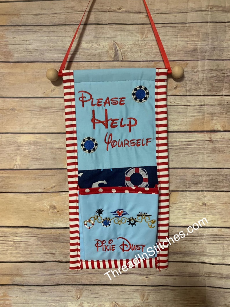 Op de afbeelding: Een lichtblauwe stoffen wandhanger met rood-witte strepen. De tekst "PLEASE HELP YOURSELF" is rood geborduurd. Extra geborduurde details zijn nautische elementen en de woorden "Pixie Dust".
