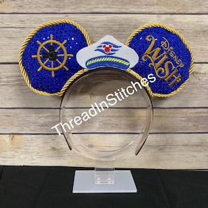 May include: Blue sequined mouse ears with gold trim and a ship's wheel design. The right ear features the text "Disney Wish" in gold. A white and blue captain's hat is centered on the headband. The text "ThreadInStitches" is printed on the headband.
