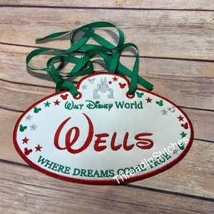 May include: White oval-shaped name tag with red and green embroidery. The tag says "Walt Disney World" and "Wells" in red. The tag also says "Where Dreams Come True" in green. The tag has a green ribbon attached.