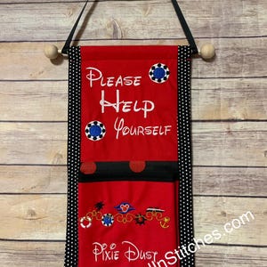 May include: Red fabric wall hanging with the text "Please Help Yourself" and "Pixie Dust" in white script. The banner has black polka dot trim and embroidered details, including blue and white circular designs and Disney-themed imagery.