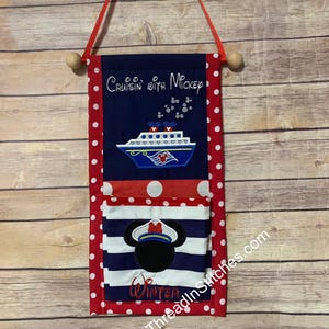 May include: A fabric wall hanging with a red polka dot border and a navy blue top section that reads "Cruisin' with Mickey". Below is a cruise ship design and a pocket with a Mickey Mouse captain design. The word "Winter" is embroidered on the pocket.