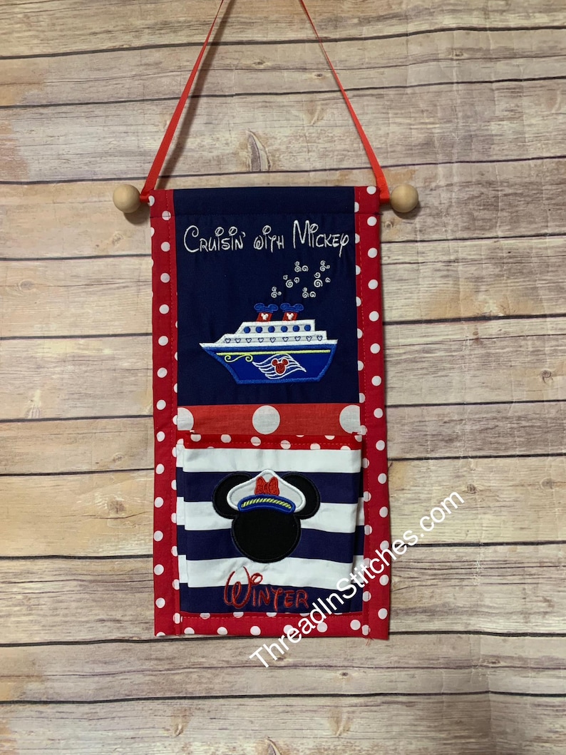 May include: A fabric wall hanging with a red polka dot border and a navy blue top section that reads "Cruisin' with Mickey". Below is a cruise ship design and a pocket with a Mickey Mouse captain design. The word "Winter" is embroidered on the pocket.