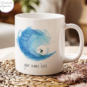 Può includere: Tazza in ceramica bianca con un'illustrazione ad acquerello di un'onda blu e un surfista. Il testo "your name here" è stampato sulla tazza.