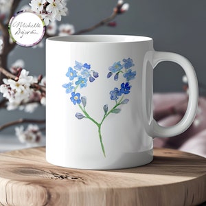 Op de afbeelding: Witte keramische mok met een aquarelontwerp van blauwe vergeet-mij-nietjes in een hartvorm. De mok heeft een gebogen oor en de handtekening van de kunstenaar, "Michelle Dujardin", staat in een cirkel aan de zijkant.