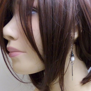 May include: A close-up of a mannequin wearing a pair of dangle earrings. The earrings feature a combination of black, white, and silver elements. The design includes small beads and a long, thin, silver bar that hangs down.