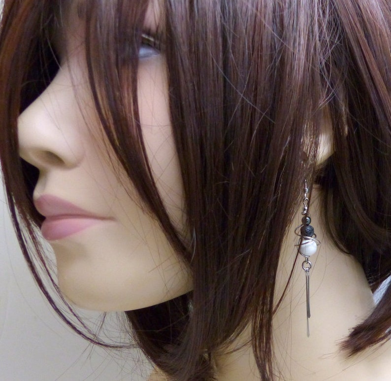 May include: A close-up of a mannequin wearing a pair of dangle earrings. The earrings feature a combination of black, white, and silver elements. The design includes small beads and a long, thin, silver bar that hangs down.
