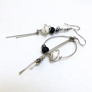 May include: A pair of handmade dangle earrings. Each earring features a silver-tone metal design with white, black, and gray beads. The earrings have a unique, abstract shape with a hook closure.