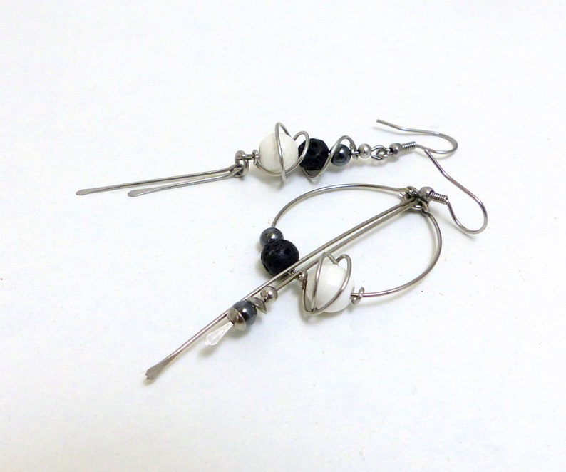 May include: A pair of handmade dangle earrings. Each earring features a silver-tone metal design with white, black, and gray beads. The earrings have a unique, abstract shape with a hook closure.