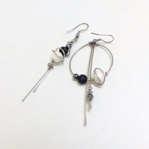 May include: A pair of asymmetrical earrings with a modern design. One earring features a series of black and white beads with dangling silver rods. The other earring has a circular design with black and white beads and silver rods.