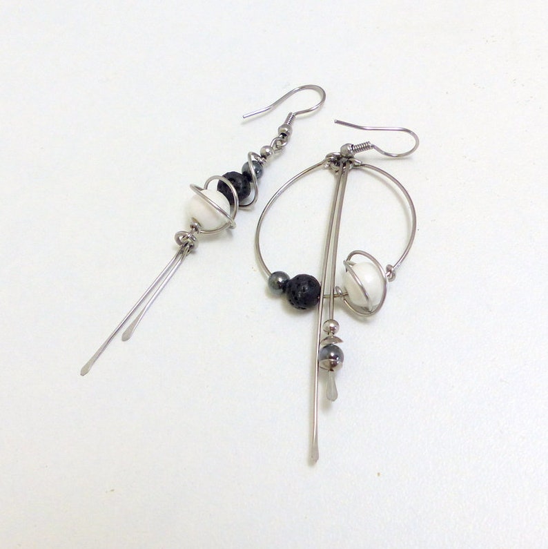 May include: A pair of asymmetrical earrings with a modern design. One earring features a series of black and white beads with dangling silver rods. The other earring has a circular design with black and white beads and silver rods.