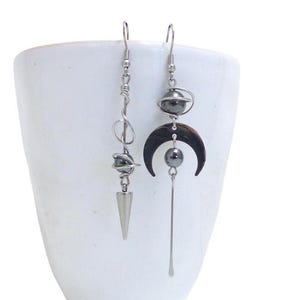 May include: A pair of silver earrings with black hematite beads and geometric shapes. One earring features a crescent moon shape and the other has a spiral design with a pointed tip.