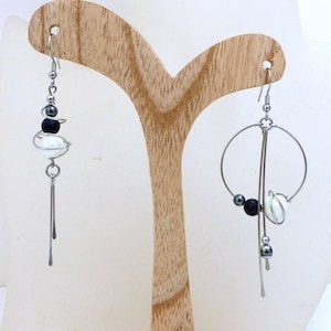 May include: Silver-tone dangle earrings with a modern design. Each earring features a hook, a series of beads, and a long, thin bar. One earring has a circular design. The beads are black, white, and silver.
