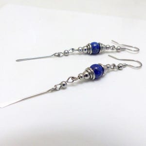 Peut inclure: Une paire de boucles d'oreilles pendantes avec une perle de gemme bleue et des détails en métal argenté. Les boucles d'oreilles ont une longue barre argentée et un crochet d'oreille.