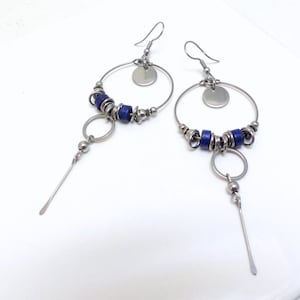 Op de afbeelding: Een paar zilverkleurige oorringen met donkerblauwe kralen en hangende zilveren accenten. Elke oorbel heeft een cirkelvormig ontwerp met een kleine, platte schijf aan de bovenkant en een lange, slanke staaf eronder. Op een witte achtergrond.