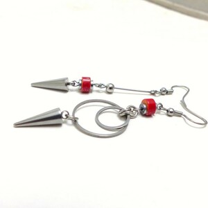 Peut inclure: Une paire de boucles d'oreilles argentées avec une perle rouge et un motif circulaire. Les boucles d'oreilles sont en métal et ont une pointe acérée en haut.